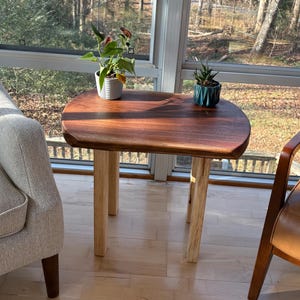 May include: A small, handcrafted wooden side table with a rich, dark brown tabletop and light-colored wooden legs. The table has a smooth, rounded shape and is positioned near a window. Two small potted plants sit on the table.