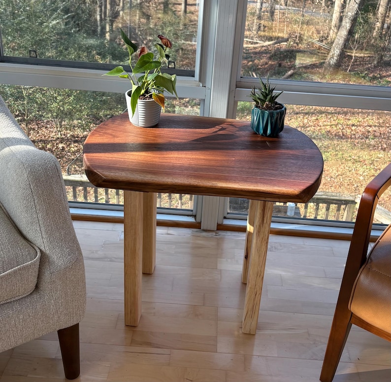 May include: A small, handcrafted wooden side table with a rich, dark brown tabletop and light-colored wooden legs. The table has a smooth, rounded shape and is positioned near a window. Two small potted plants sit on the table.