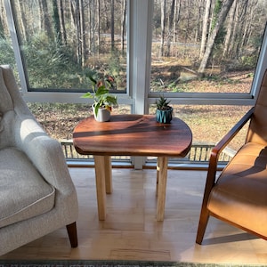 May include: A small, wooden side table with a dark brown tabletop and light-colored legs. The table is placed between two chairs in a sunroom, with plants on top. The table is approximately 24 inches long and 18 inches wide.