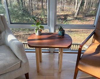 May include: A small, wooden side table with a dark brown tabletop and light-colored legs. The table is placed between two chairs in a sunroom, with plants on top. The table is approximately 24 inches long and 18 inches wide.