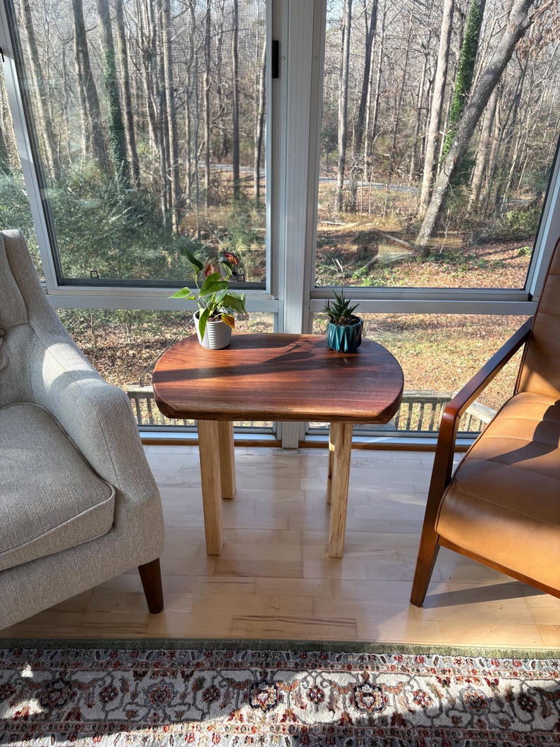 May include: A small, wooden side table with a dark brown tabletop and light-colored legs. The table is placed between two chairs in a sunroom, with plants on top. The table is approximately 24 inches long and 18 inches wide.