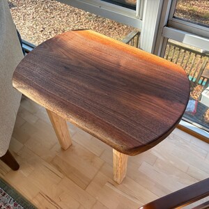 May include: A small, handcrafted wooden side table with a dark brown, live-edge top and light-colored legs. The table is made of solid wood and has a natural finish. The table is approximately 24 inches long, 16 inches wide, and 18 inches tall.