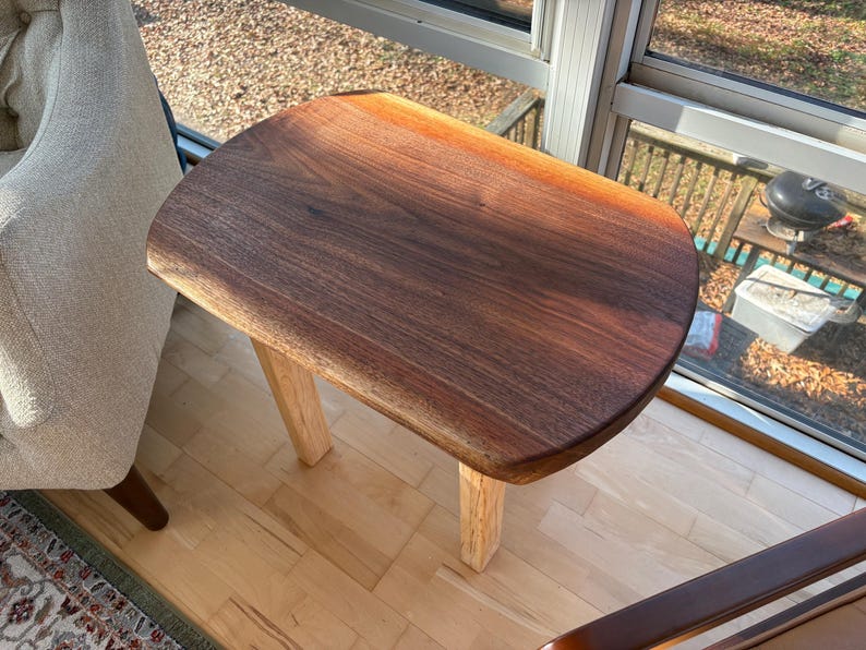 May include: A small, handcrafted wooden side table with a dark brown, live-edge top and light-colored legs. The table is made of solid wood and has a natural finish. The table is approximately 24 inches long, 16 inches wide, and 18 inches tall.