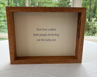 May include: A framed print with the quote "true love is when both people think they are the lucky one." The frame is made of light brown wood, and the background is a neutral off-white color. The print is displayed on a white surface.