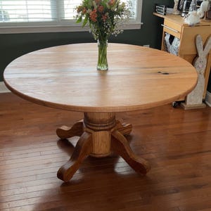 May include: A round wooden dining table with a pedestal base. The table is made of light-colored wood and has a smooth surface. A vase of flowers sits in the center of the table. The table is in a well-lit room.