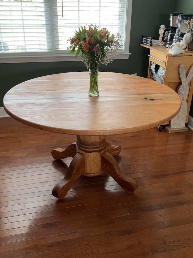 May include: A round wooden dining table with a pedestal base. The table is made of light-colored wood and has a smooth surface. A vase of flowers sits in the center of the table. The table is in a well-lit room.