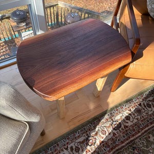 May include: A small, dark brown wooden side table with a rounded top and light-colored legs. The table is positioned next to a brown leather chair and a light-colored upholstered armchair. The table is approximately 24 inches long.
