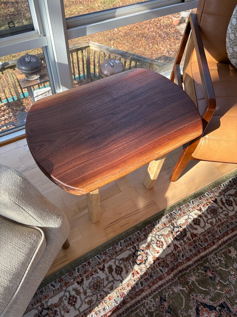 May include: A small, dark brown wooden side table with a rounded top and light-colored legs. The table is positioned next to a brown leather chair and a light-colored upholstered armchair. The table is approximately 24 inches long.
