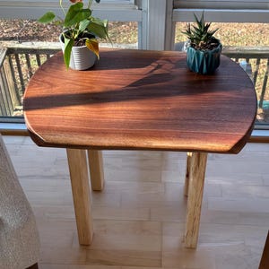 May include: A small, handcrafted wooden side table with a rich, dark brown tabletop and light-colored wooden legs. The table has a smooth, rounded shape and is topped with two potted plants. The table is placed near a window.