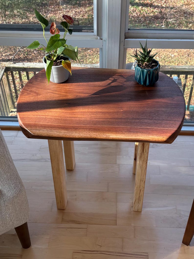 May include: A small, handcrafted wooden side table with a rich, dark brown tabletop and light-colored wooden legs. The table has a smooth, rounded shape and is topped with two potted plants. The table is placed near a window.