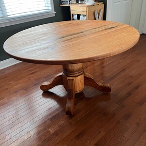 May include: A round wooden dining table with a light brown finish. The table has a pedestal base with four curved legs. The tabletop is approximately 48 inches in diameter. The table is set on a hardwood floor.