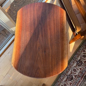 May include: A solid wood side table with a dark brown finish. The table has a rounded rectangular top and a cylindrical body. The wood grain is visible. The table is approximately 24 inches tall.