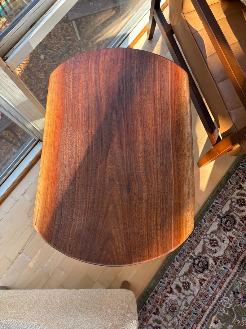 May include: A solid wood side table with a dark brown finish. The table has a rounded rectangular top and a cylindrical body. The wood grain is visible. The table is approximately 24 inches tall.