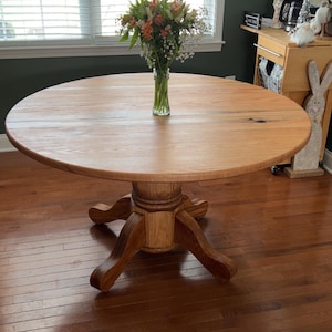 May include: A round wooden dining table with a pedestal base. The tabletop is light brown, and the base is a darker shade of brown. A vase of flowers sits in the center. The table is approximately 48 inches in diameter.