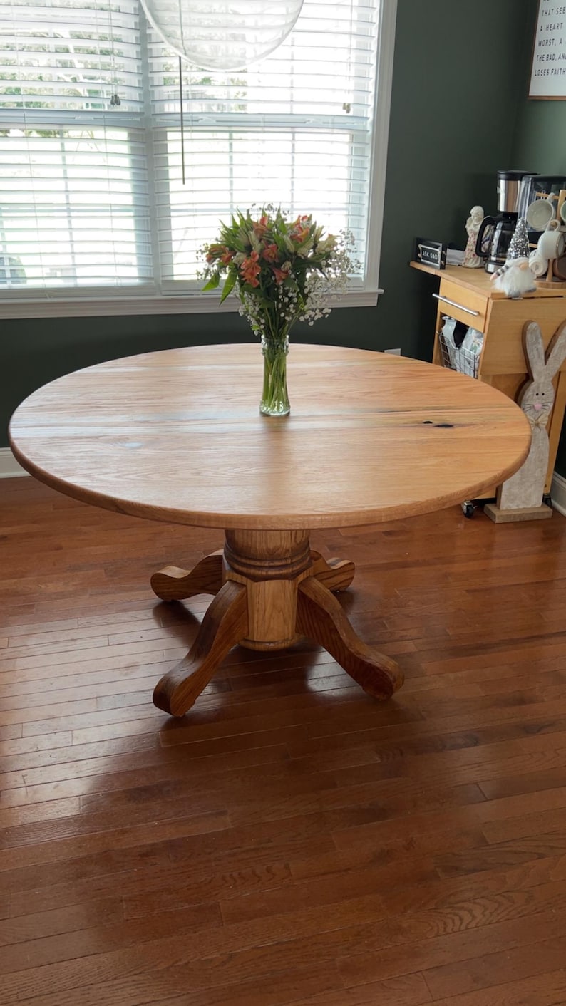 May include: A round wooden dining table with a pedestal base. The tabletop is light brown, and the base is a darker shade of brown. A vase of flowers sits in the center. The table is approximately 48 inches in diameter.