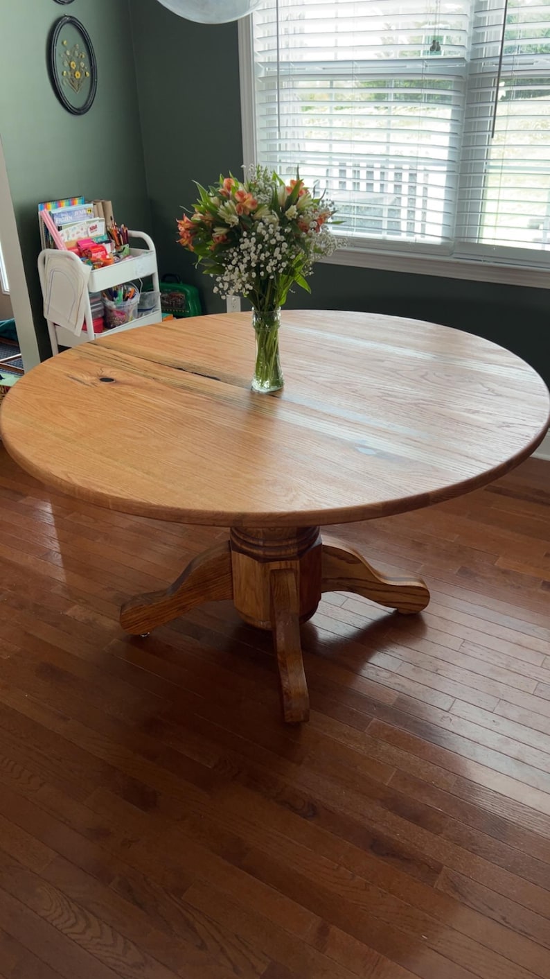 May include: A round wooden dining table with a light brown finish. The table has a pedestal base and is approximately 48 inches in diameter. A vase of flowers sits on the table. The table is in a room with hardwood floors and a window.