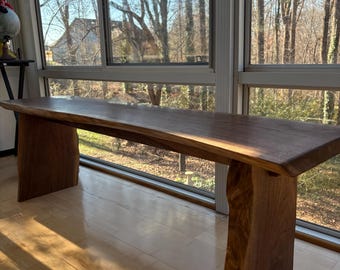May include: A long, rectangular wooden bench featuring a smooth, dark brown surface with a natural live edge. The bench is supported by two thick, solid wooden slab legs that mirror the organic, textured edge of the seat, creating a sturdy structure.