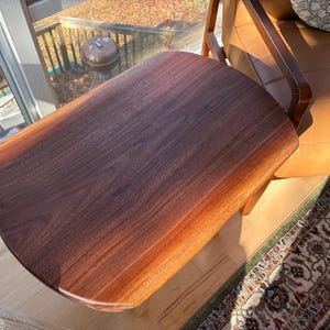 May include: A solid wood side table with a dark brown finish. The table has a rounded edge and is attached to a wooden frame. The table is next to a brown leather chair and a patterned rug.