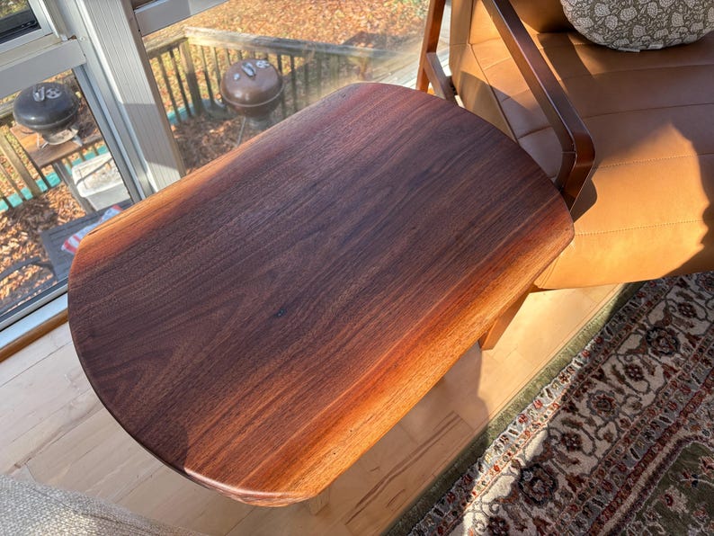 May include: A solid wood side table with a dark brown finish. The table has a rounded edge and is attached to a wooden frame. The table is next to a brown leather chair and a patterned rug.