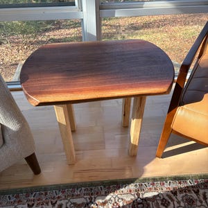 May include: A small, handcrafted wooden side table with a dark brown, oval-shaped top and four light-colored, square legs. The table is made of natural wood and has a smooth, polished finish. The table is approximately 24 inches long, 18 inches wide, and 20 inches tall.