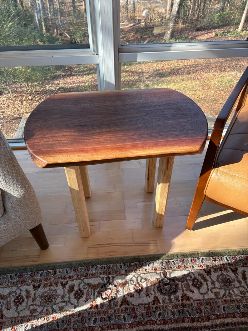 May include: A small, handcrafted wooden side table with a dark brown, oval-shaped top and four light-colored, square legs. The table is made of natural wood and has a smooth, polished finish. The table is approximately 24 inches long, 18 inches wide, and 20 inches tall.