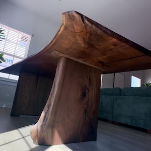 May include: A large, solid wood table with a natural, live edge design. The table has a rich, dark brown color and is supported by two thick, angled legs. The tabletop is approximately 120 inches long and 40 inches wide, with a unique, curved shape.