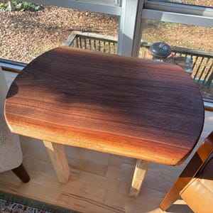 May include: A small, oval-shaped wooden side table with a dark brown, polished top and light-colored legs. The table is made of solid wood and has a natural wood grain. The table is approximately 24 inches long.