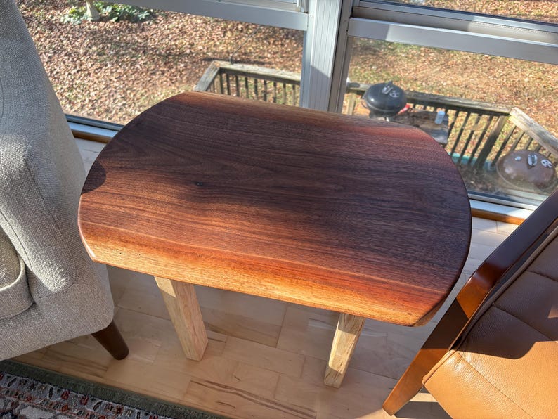 May include: A small, oval-shaped wooden side table with a dark brown, polished top and light-colored legs. The table is made of solid wood and has a natural wood grain. The table is approximately 24 inches long.