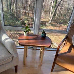 May include: A small, wooden side table with a dark brown, rounded top and light-colored legs. Two small potted plants sit on the table. The table is in front of a large window with a view of trees.
