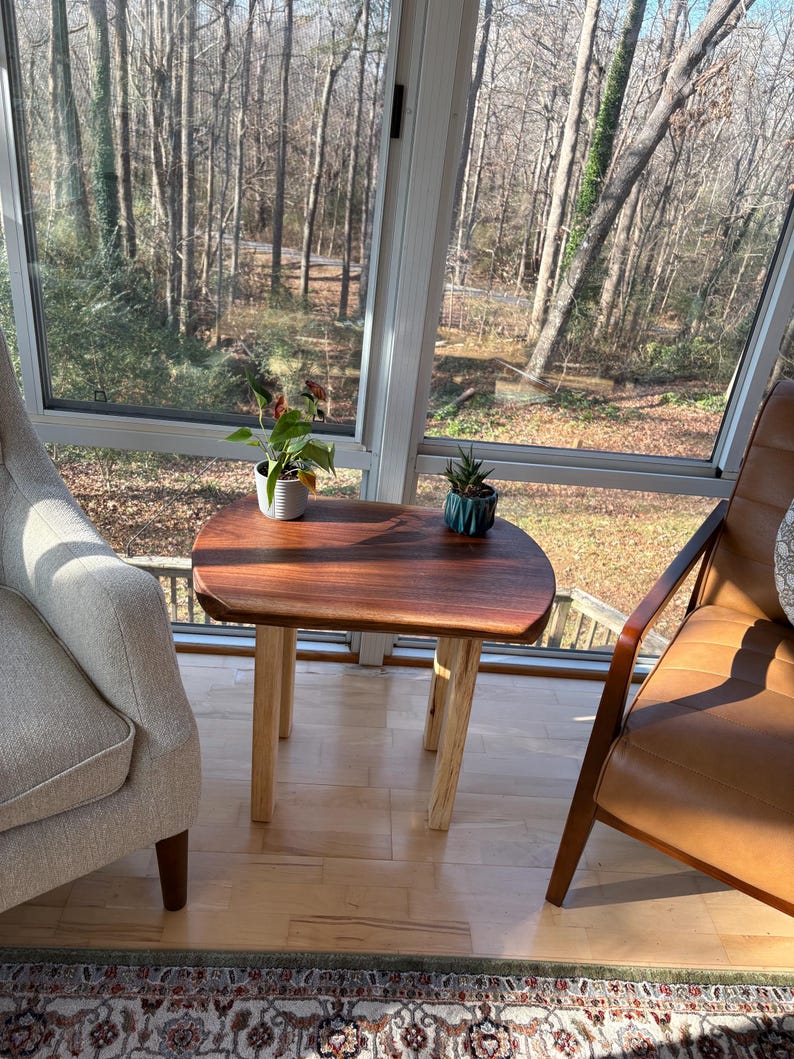 May include: A small, wooden side table with a dark brown, rounded top and light-colored legs. Two small potted plants sit on the table. The table is in front of a large window with a view of trees.