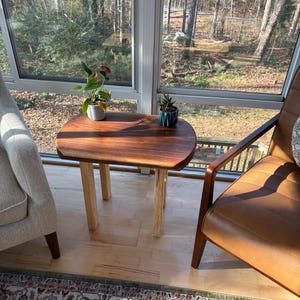 May include: A small, wooden side table with a dark brown, oval-shaped top and light-colored square legs. The table is placed between a beige armchair and a brown leather chair, with plants on top. The table is approximately 24 inches tall.