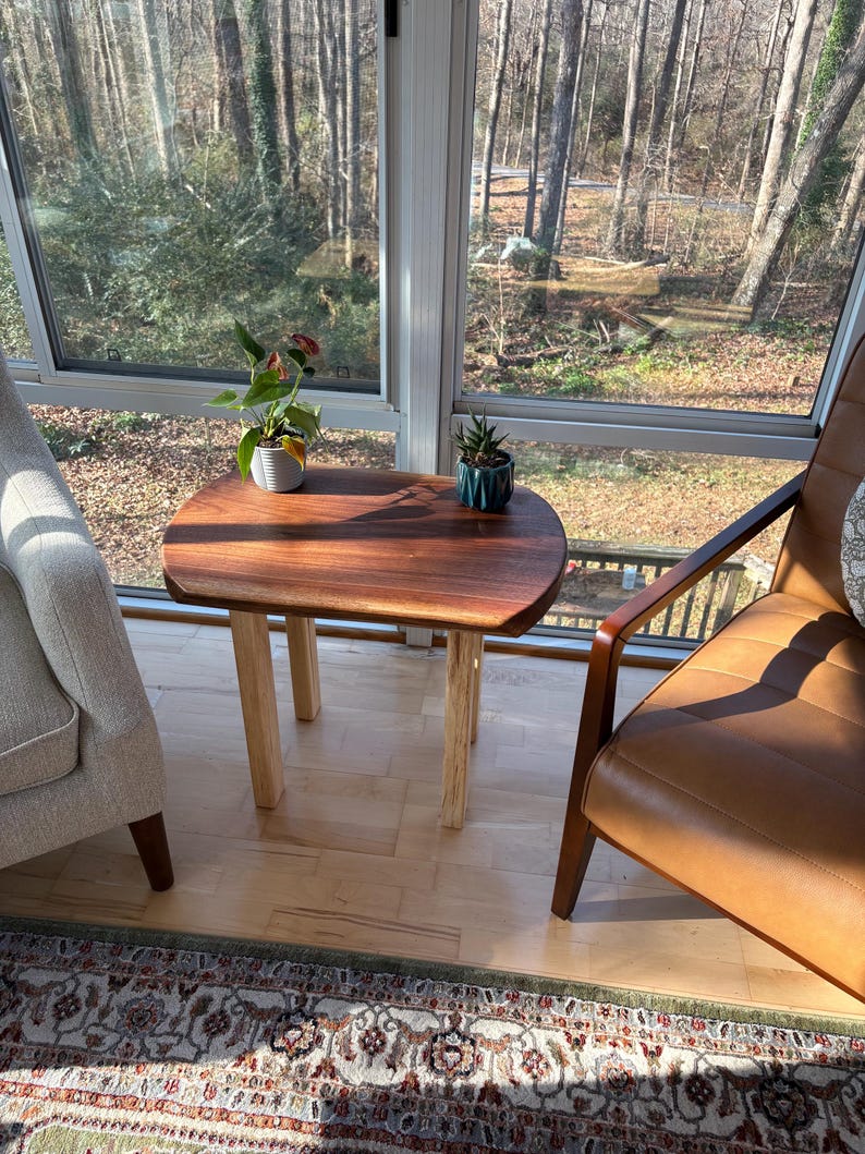 May include: A small, wooden side table with a dark brown, oval-shaped top and light-colored square legs. The table is placed between a beige armchair and a brown leather chair, with plants on top. The table is approximately 24 inches tall.