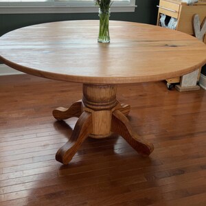 May include: A round wooden dining table with a pedestal base. The table is made of light-colored wood and has a smooth surface. A vase of flowers sits in the center. The table is approximately 48 inches in diameter.
