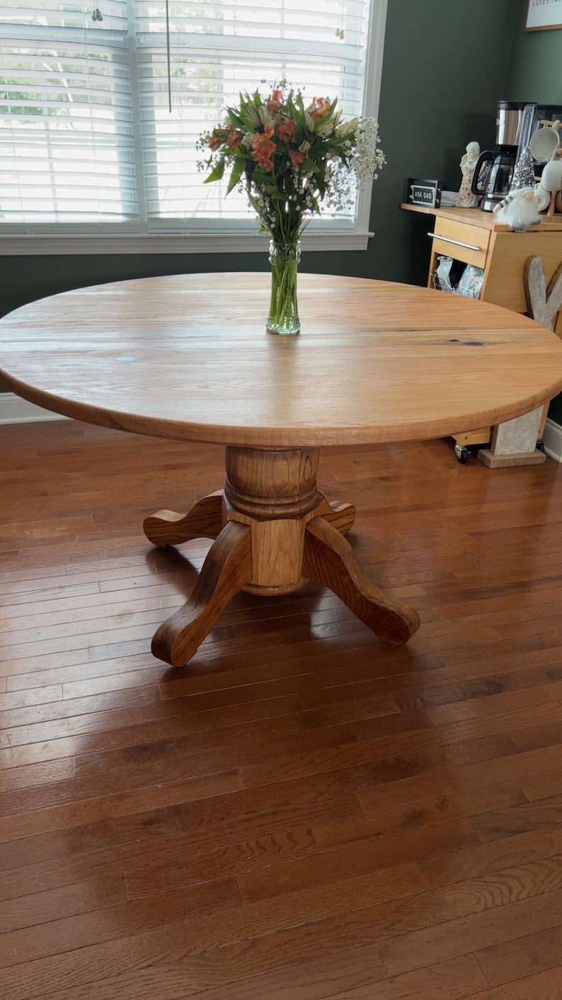 May include: A round wooden dining table with a pedestal base. The table is made of light-colored wood and has a smooth surface. A vase of flowers sits in the center. The table is approximately 48 inches in diameter.