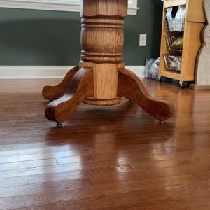 May include: A round wooden dining table with a pedestal base. The table has a light brown finish and is supported by a carved pedestal and four curved feet. The table is set on a hardwood floor.