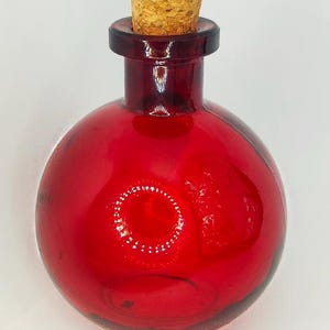 Puede incluir: Una botella de vidrio rojo con un tapón de corcho. La botella tiene forma redonda con un cuello corto. El corcho es de color marrón claro. El texto "BOTTLES TOPS & CANDLES" está en la parte inferior de la imagen.