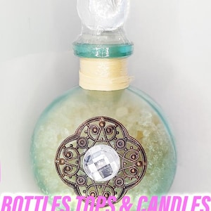 Puede incluir: Una botella de vidrio decorativa con un tapón transparente y un cuello turquesa. La botella presenta un diseño de filigrana de bronce con un gran cristal facetado en el centro. El texto "BOTTLES TOPS & CANDLES" está en la parte inferior.