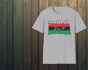 May include: A gray t-shirt with the word "juneteenth" in white script above a flag design in red, black, and green. The text "June 19th" is below the flag. The shirt is displayed against a wooden background.