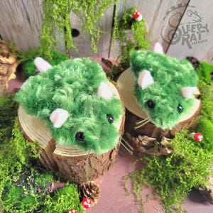 May include: Two plush green triceratops toys with white horns and ears, and black eyes, rest on wooden slices surrounded by moss and small red and white mushroom decorations. The toys are part of the Sleepy Rat Archives collection.