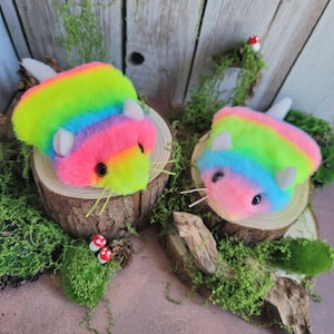 May include: Two plush toys shaped like small rodents, covered in vibrant rainbow-colored faux fur. Each toy features white ears, black bead eyes, and thin whiskers. They are positioned on circular wooden slices decorated with green moss.