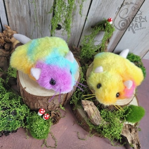 May include: Two plush rodents with soft, tie-dye fur in shades of yellow, blue, and pink. Each plush features white ears, black eyes, and thin whiskers. The rodents are positioned on circular wooden slices decorated with green moss.