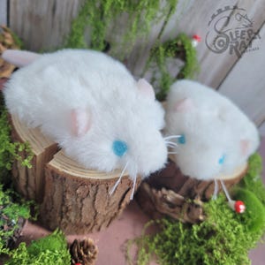 May include: Two plush white mice with pink ears and blue eyes rest on a wooden surface. The mice have long white whiskers and are surrounded by green moss and small red mushrooms. The Sleepy Rat Archives logo is visible in the background.