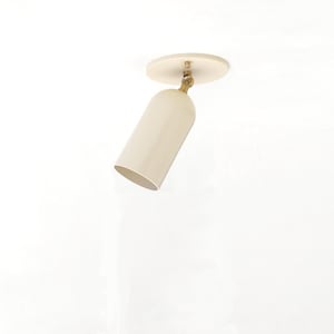 May include: A cream-colored cylindrical spotlight fixture featuring a circular ceiling mount and a brass-toned swivel joint that allows the light head to be angled. The fixture has a smooth, matte finish and a simple, functional design.