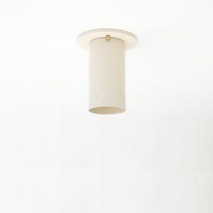 May include: A modern, cylindrical ceiling light fixture in a cream color. The light has a circular base and a cylindrical shade, with a small gold-colored accent. The fixture is suspended from the ceiling.