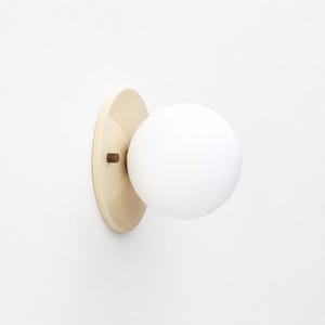 May include: A wall-mounted light fixture featuring a spherical white glass globe attached to a circular, cream-colored base. A small, dark metallic cylindrical connector joins the globe to the base, creating a clean and simple lighting design.