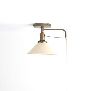 May include: A wall-mounted light fixture featuring a conical off-white metal shade attached to an antique brass finish arm. The fixture includes a circular mounting plate and a long metal pull chain for operation, extending downward from the arm.