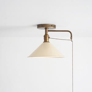 May include: A ceiling-mounted light fixture featuring a conical off-white shade and an antique brass finish. The lamp includes a horizontal arm, a circular mounting plate, and a long metal pull chain for operation, creating a functional design.