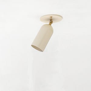 May include: A beige-colored, adjustable spotlight fixture. The lamp has a cylindrical shade and a circular base, connected by a brass-colored joint. The fixture is mounted on a white surface.