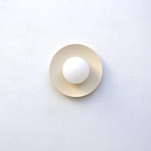 May include: A modern wall sconce featuring a white spherical light bulb centered on a circular, pale yellow disc. The sconce is mounted on a white textured wall, creating a minimalist design.