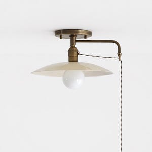 May include: A ceiling-mounted light fixture featuring a circular, off-white metal shade and a central white globe bulb. The hardware is finished in an antique brass tone, including a horizontal arm and a long, dangling metal pull chain switch.